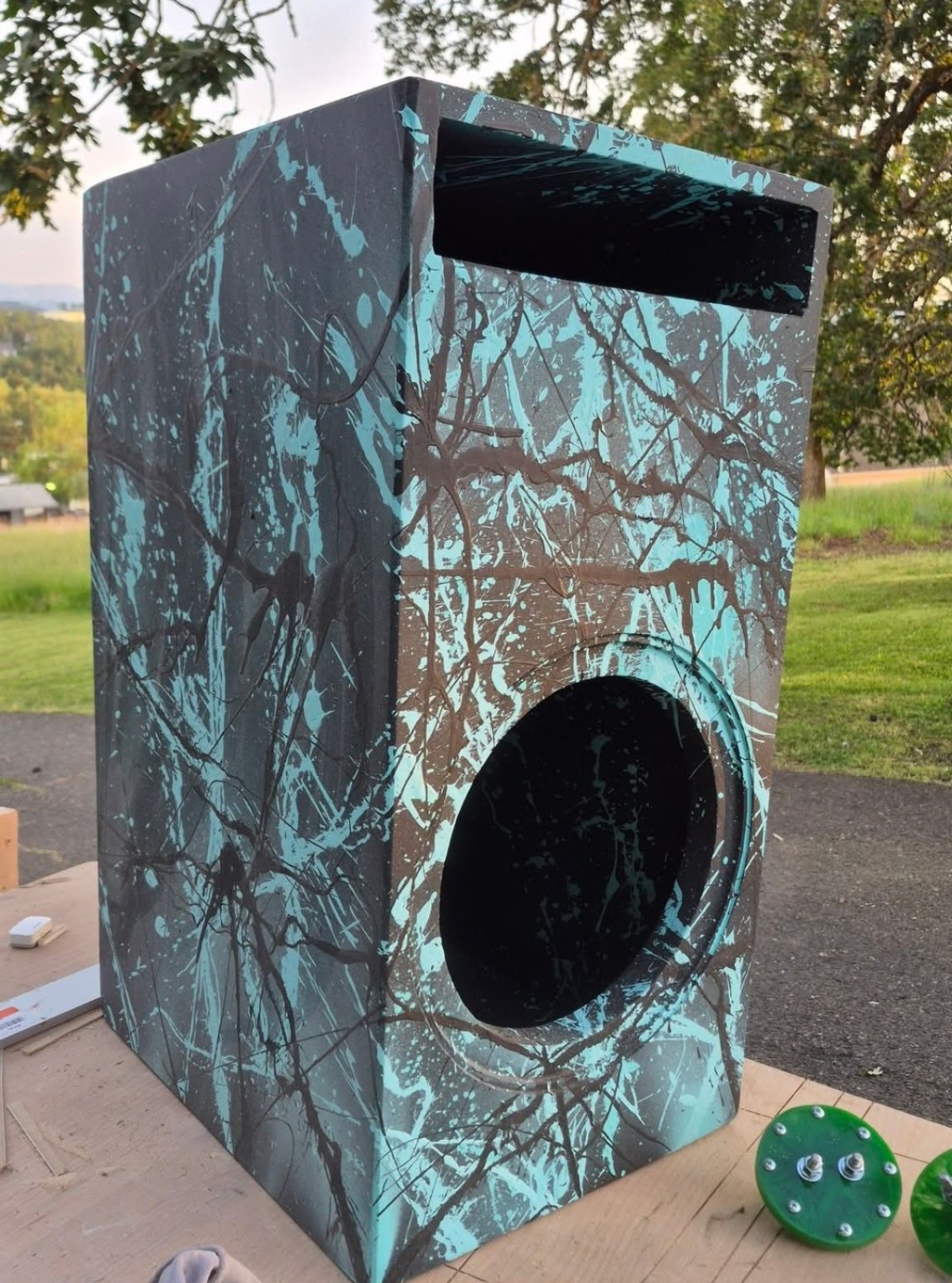 Tall turquoise and dark splatter enclosure with one round subwoofer opening and top slot port